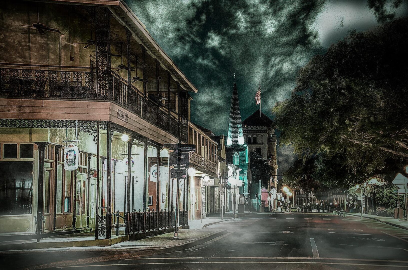 Empty street at night with iron-balcony buildings and a lit church steeple under dark clouds