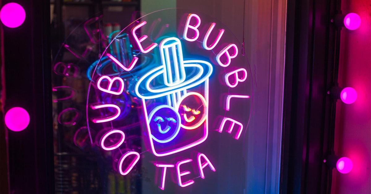 Pink and blue neon bubble tea shop sign with a cartoon cup illustration on a window