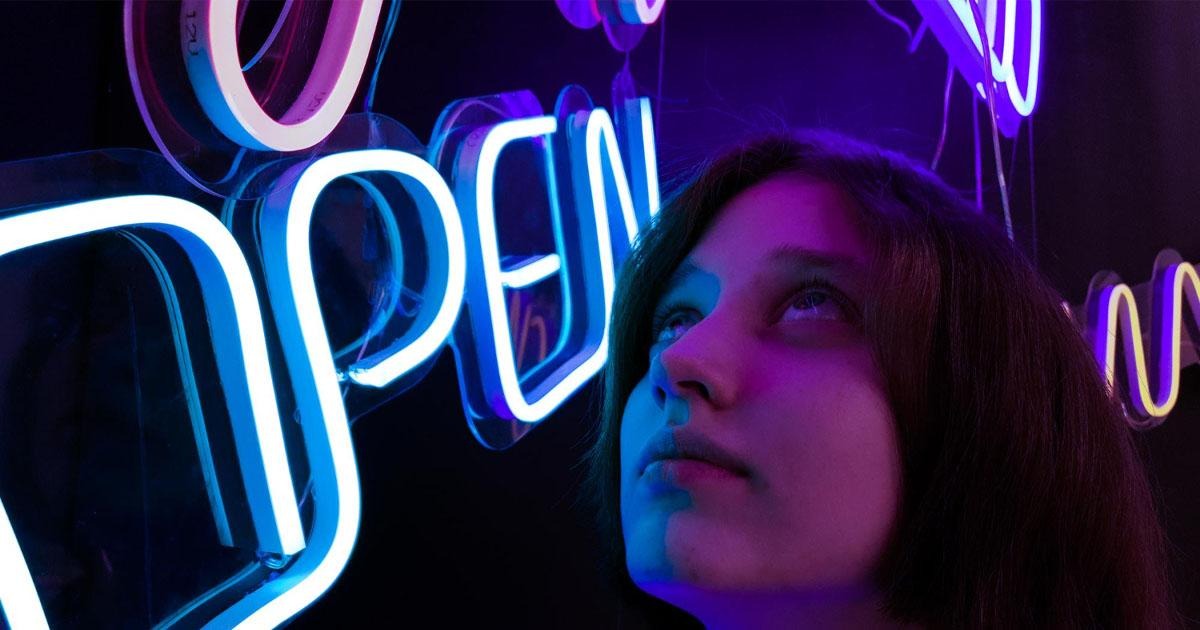 Young woman looking up at glowing blue and purple open signs in a dark room