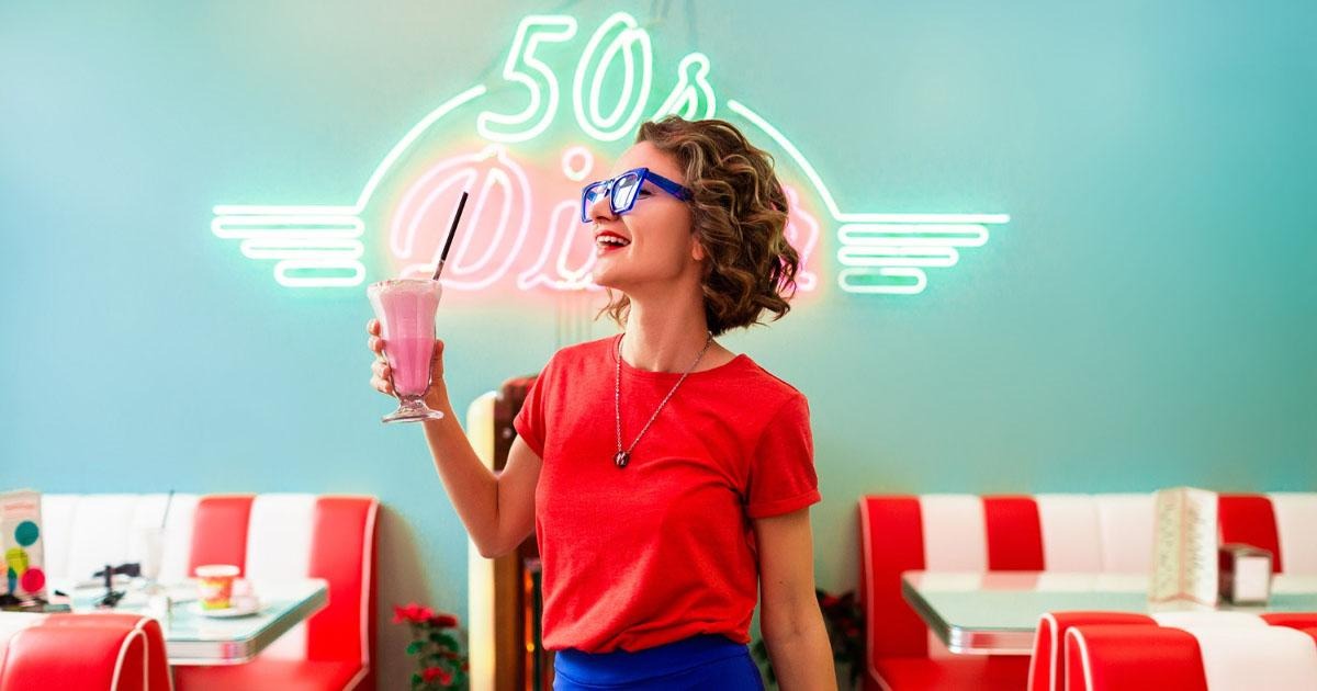Smiling woman in red top holding a pink milkshake in a retro diner with a 50s sign