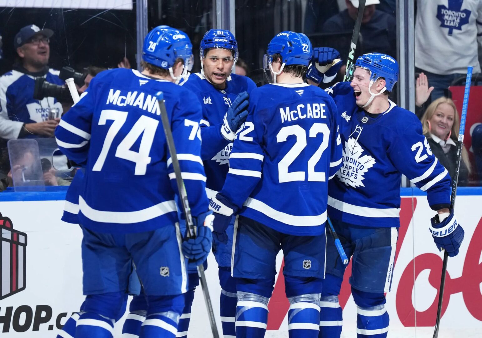 Five hockey players in blue jerseys numbered 74 and 22 celebrate near the boards as fans cheer