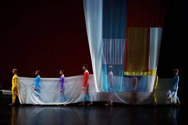 Dancers in multicolored costumes walk amid white drapes
