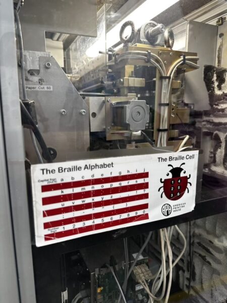 A sign showing how the Braille alphabet is laid out. A Braille printing machine is in the background. 