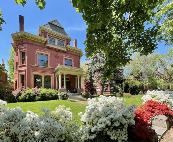 A large Victorian mansion with white and red flowers and a large green lawn in front of it. There is a clear blue sky behind it. 