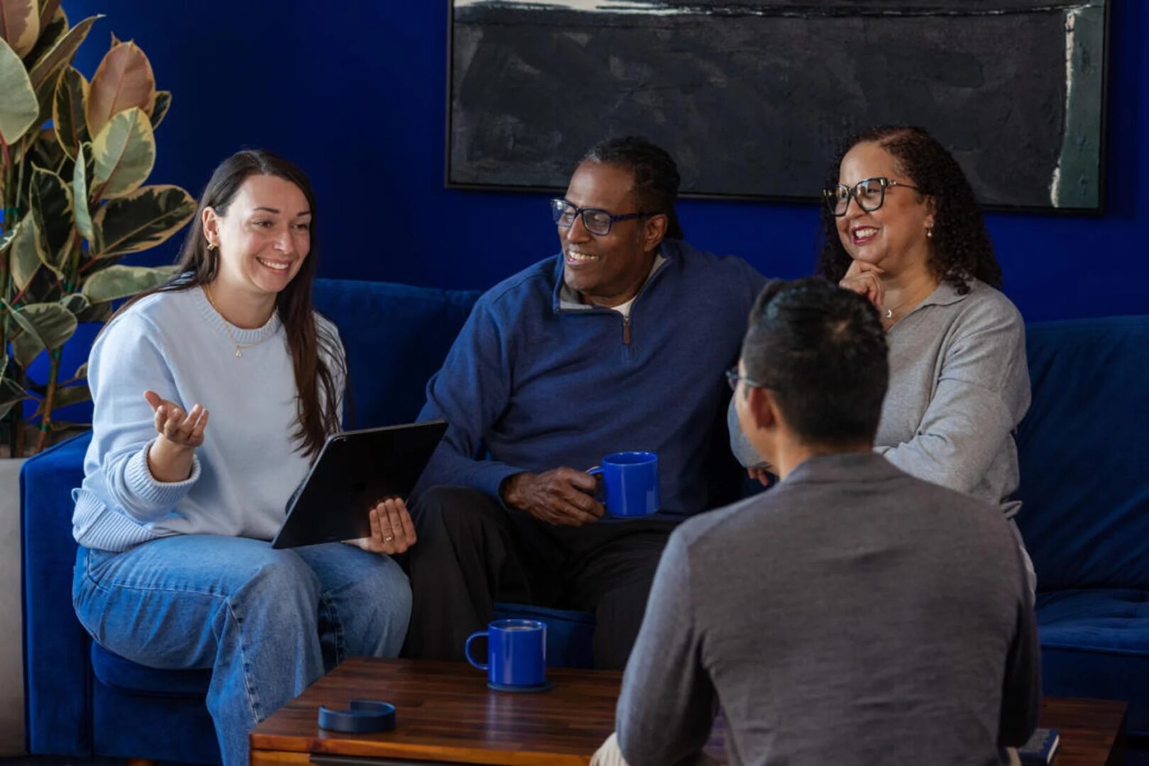 Four people chatting in a blue living room, woman holding a laptop gestures while others smile with mugs on a coffee table