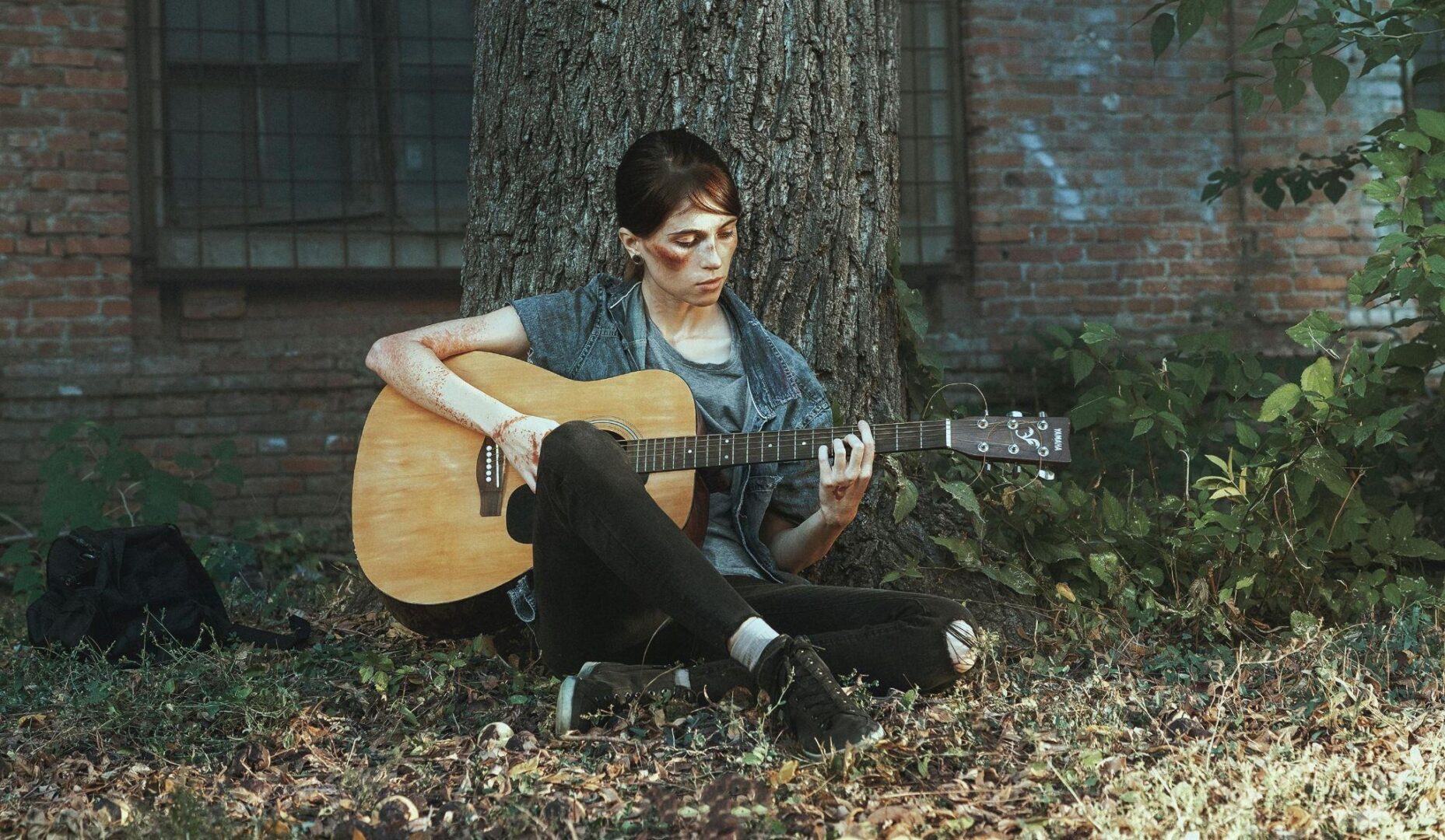 A bruised young woman sits against a tree playing an acoustic guitar outdoors near a brick building