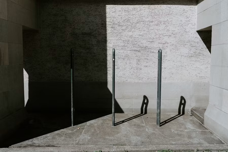 Two metal bike racks on a concrete pavement beside a textured stone wall casting sharp shadows