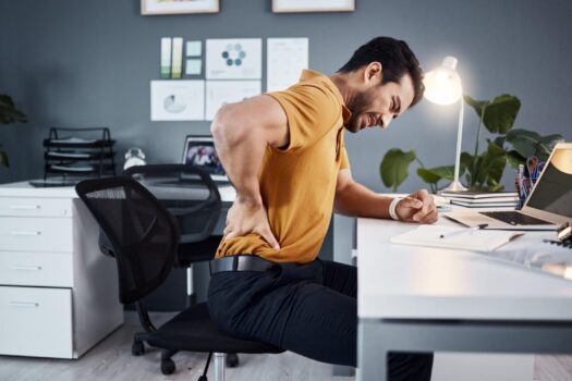 Man in orange shirt grimacing and clutching his lower back while seated at an office desk with a laptop