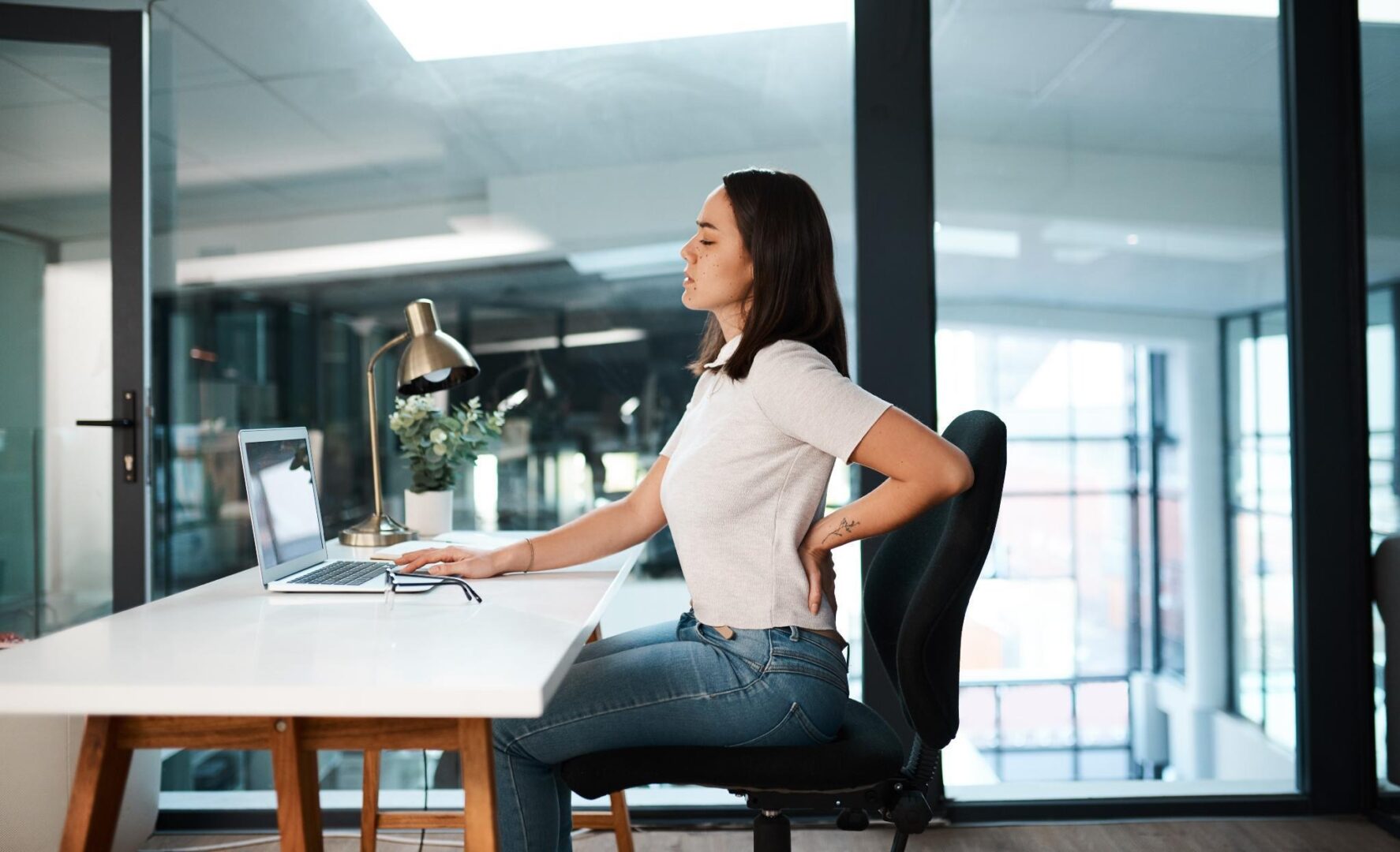 Woman in a gray top holding her lower back while sitting at a white desk with a laptop in a bright office