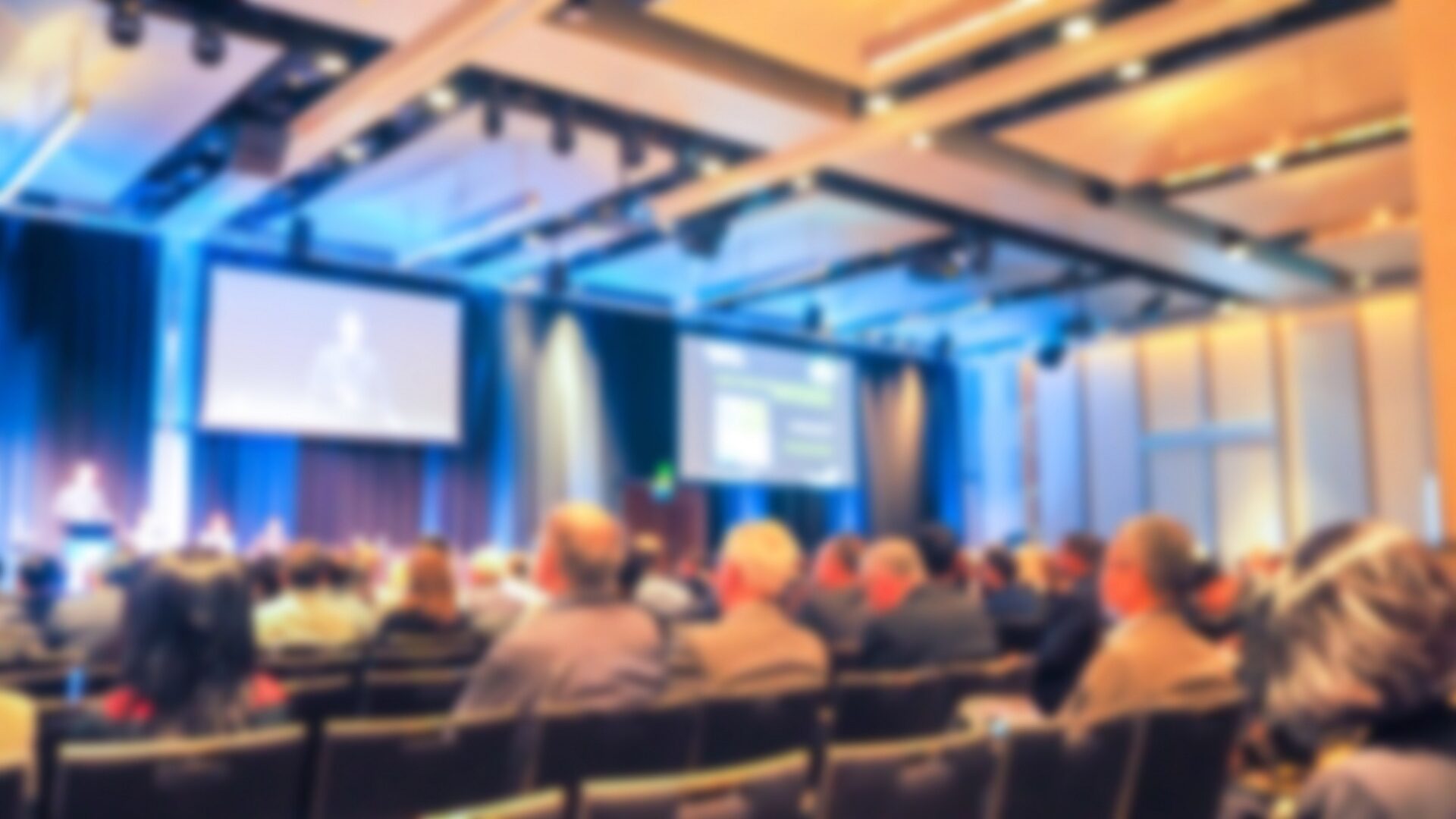 CD-Insert1 Blurred audience seated in rows facing two large projection screens in a modern hall with track lighting