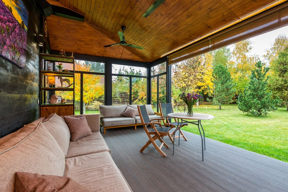 CD-Insert1 Covered patio with wood ceiling fan, beige sofa, folding chairs, small table, and green garden beyond