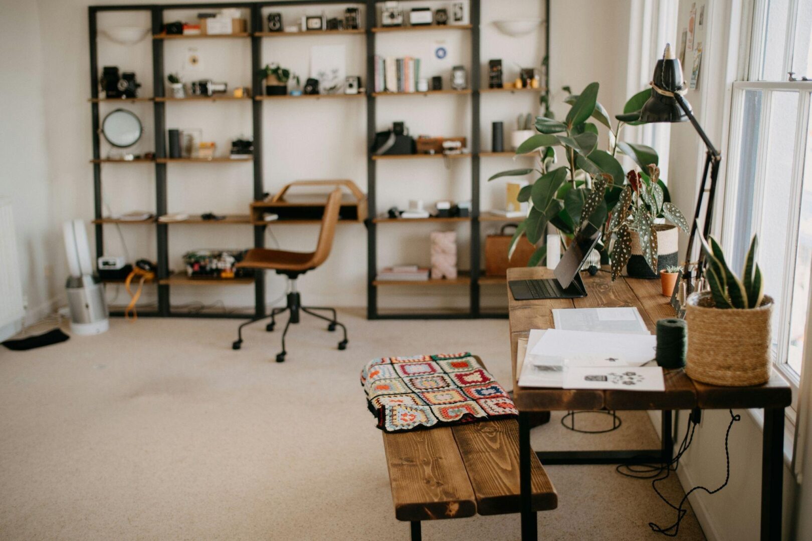 CD-Insert1 A tidy home studio with a wooden desk, laptop, plants, a desk lamp, open notebook, and shelves holding books and cameras