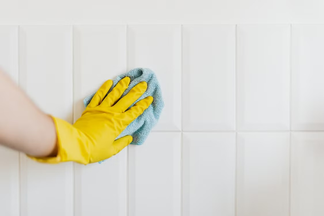 A hand in a yellow rubber glove scrubbing white wall tiles with a blue cloth