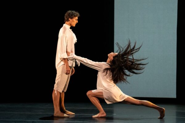A dancer in white bends before a dancer standing