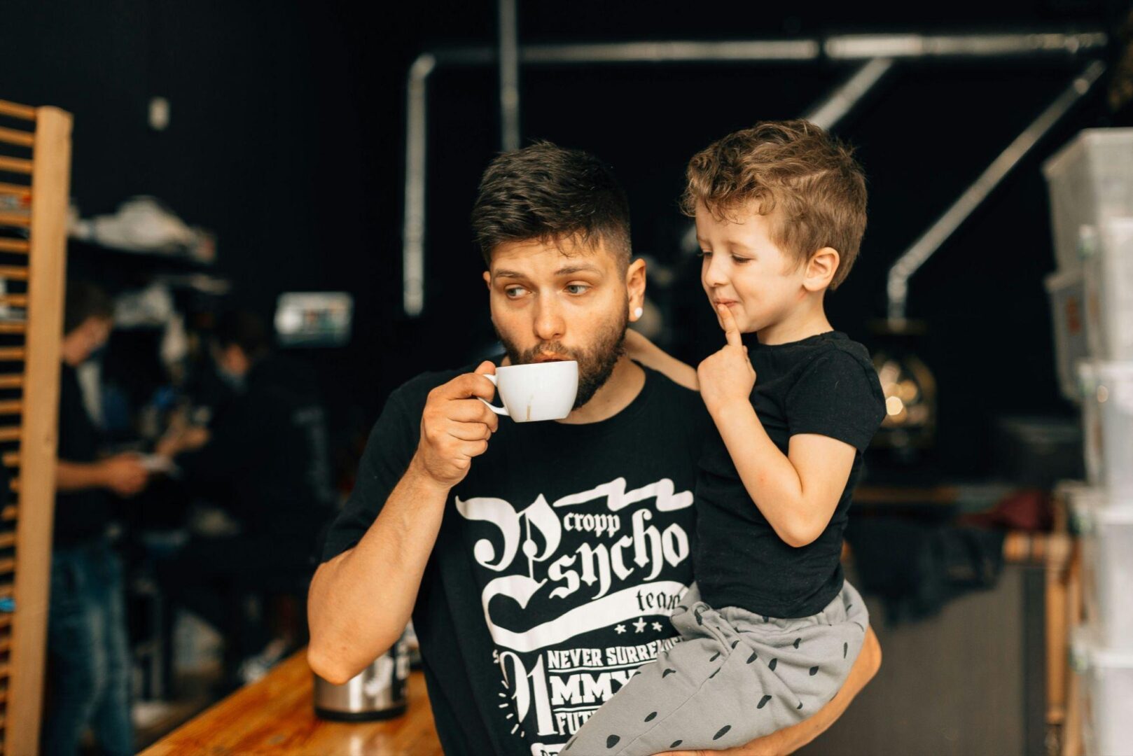 CD-Insert1 Bearded man sipping from a small white cup while holding a young boy on his arm indoors