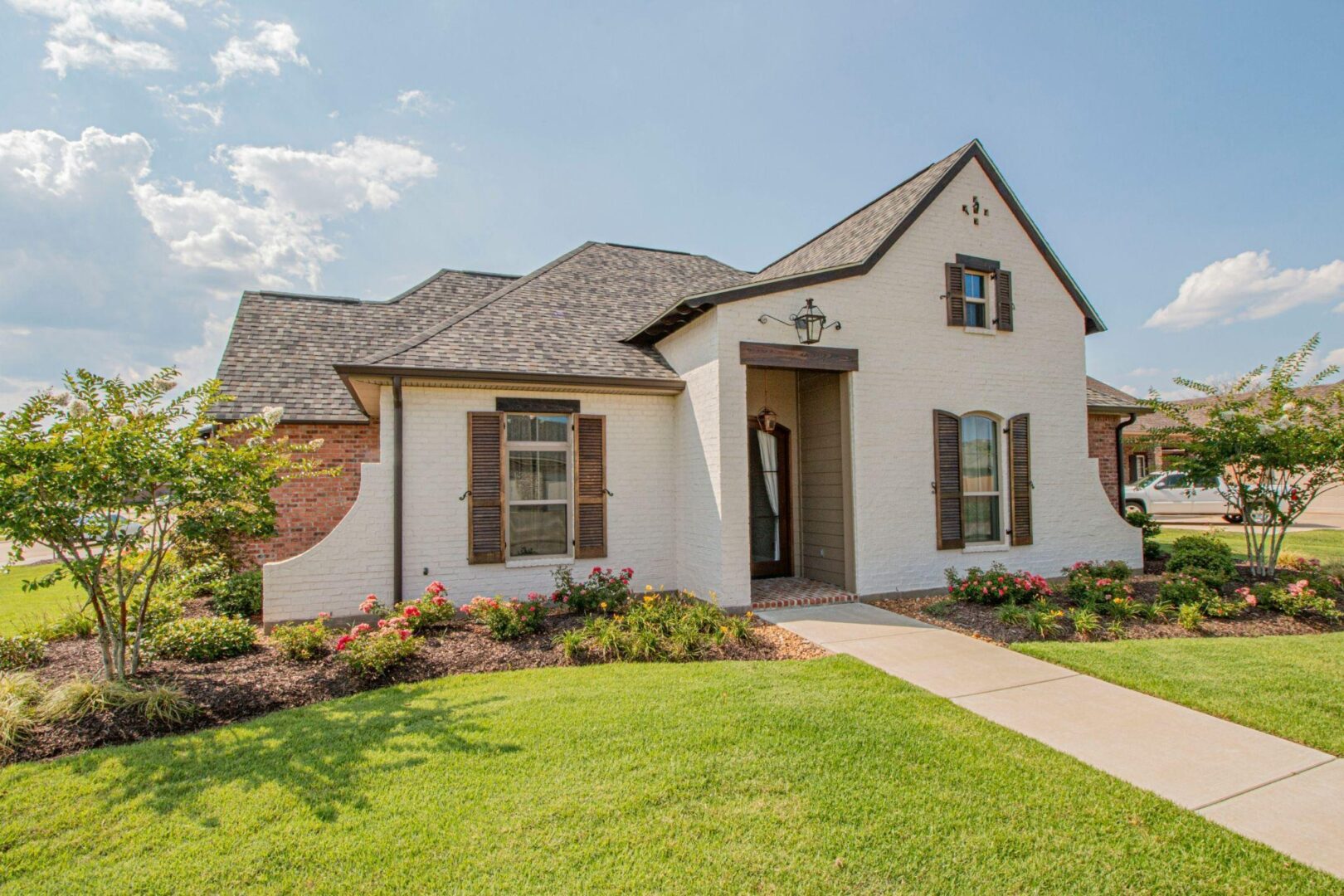 White brick house with brown shutters, manicured lawn, flower beds, and a concrete walkway leading to the front door