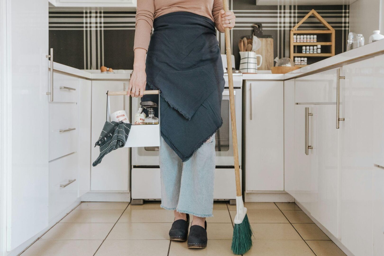 Person in apron holding a broom and small caddy with supplies standing on a tiled kitchen floor