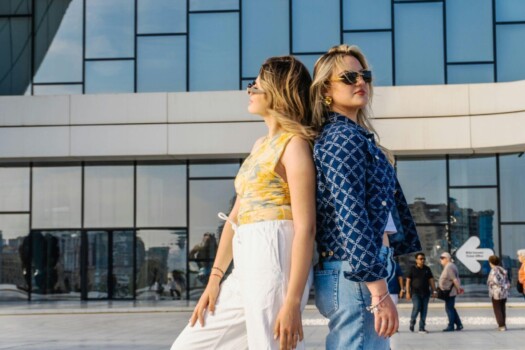 CD-Insert1 Two women in sunglasses stand back to back outdoors in front of a glass building on a sunny day