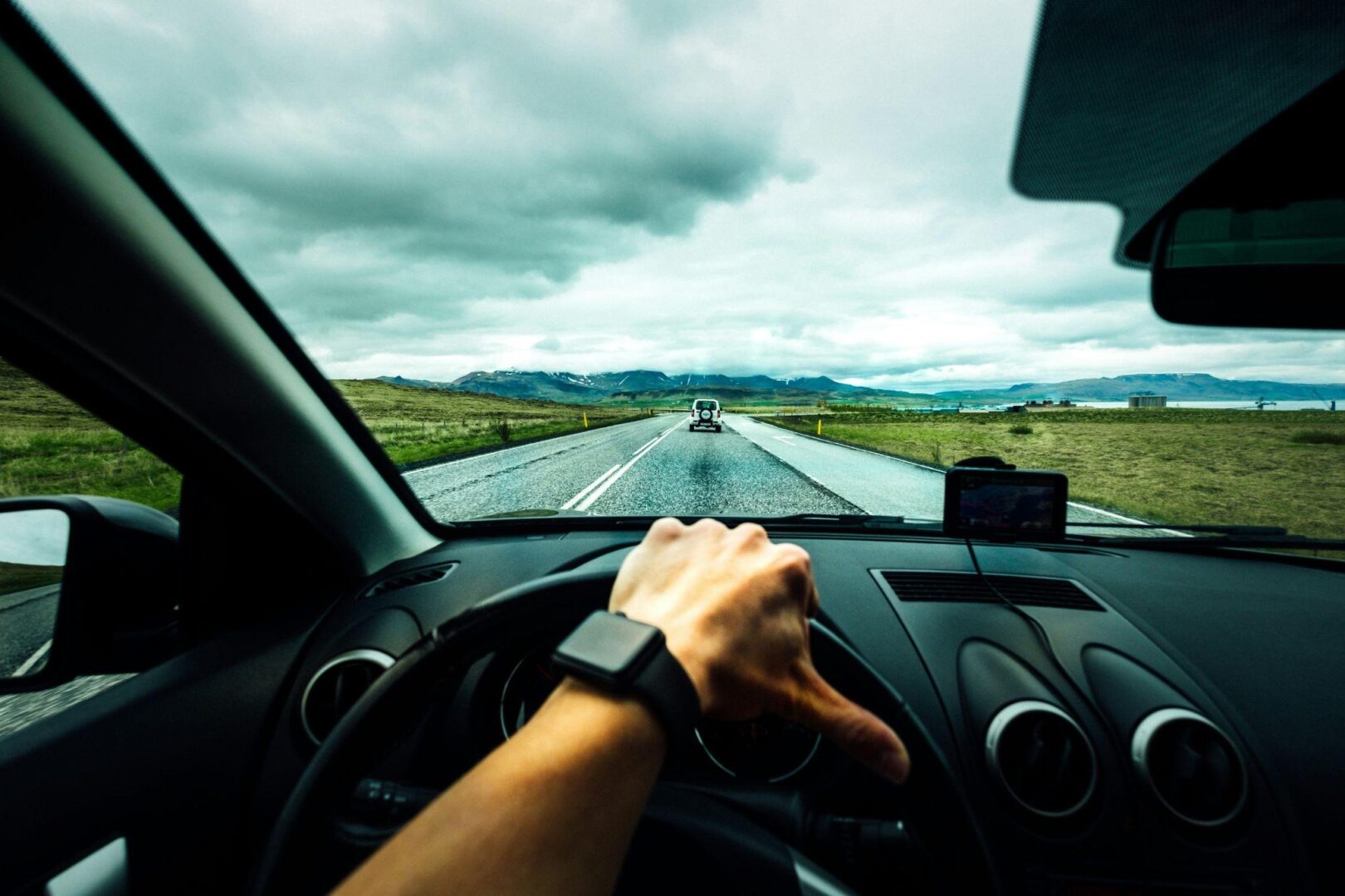 Driver's hands on steering wheel with smartwatch, dashboard with GPS device, open road ahead under cloudy skies