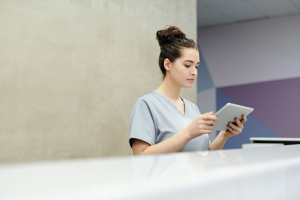 CD-healthcare-virtual-assistant-Insert1 A receptionist in scrubs using a tablet at a clinic desk