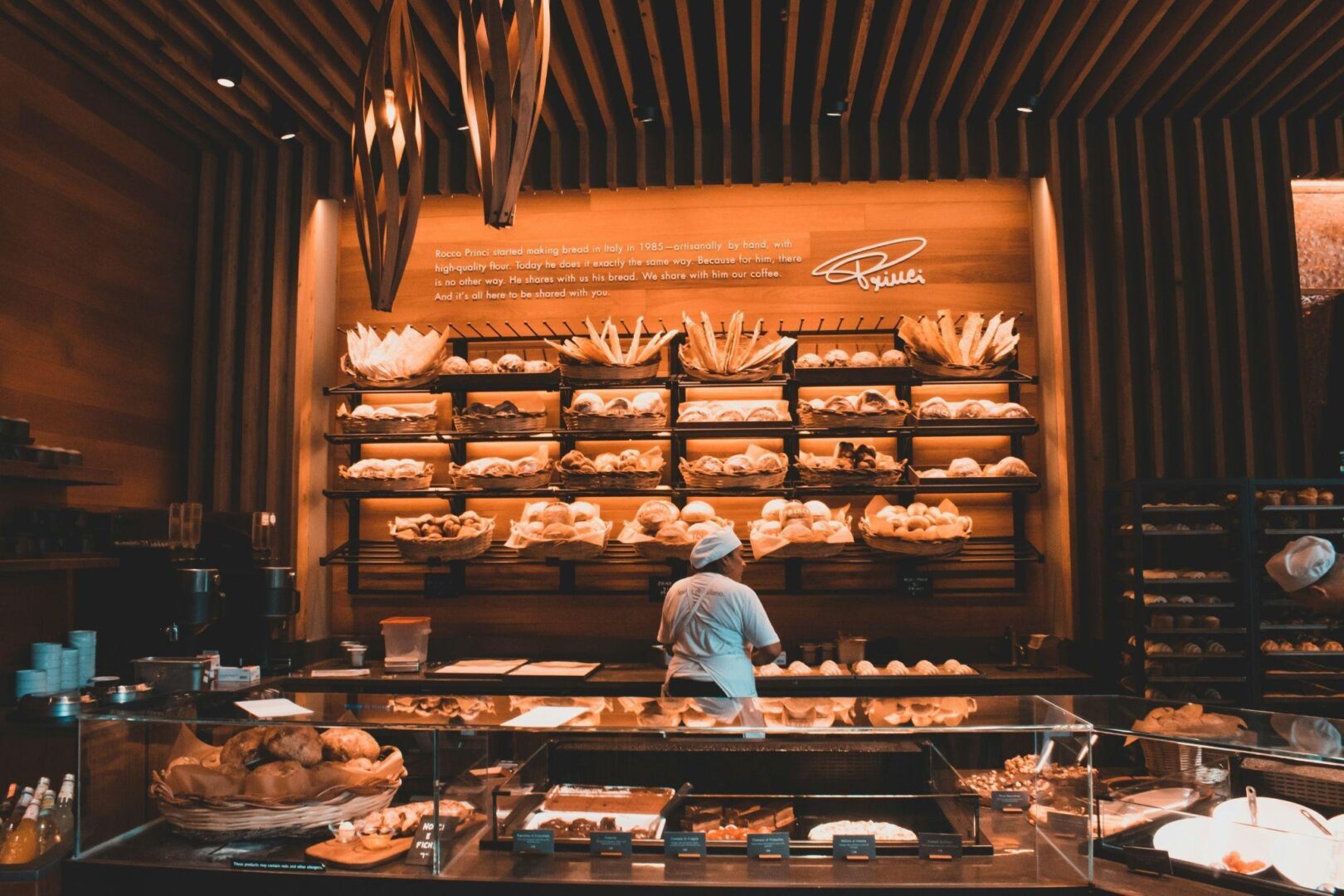 CD-Insert1 Baker in white uniform stands behind glass display counter inside warmly lit bakery with shelves of bread