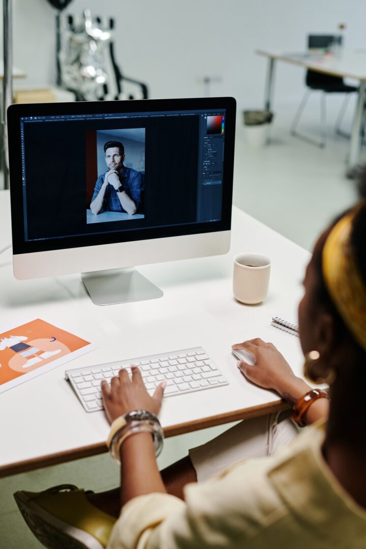 Woman at a white desk using an iMac that shows a portrait being edited, with a keyboard, mouse, and mug.