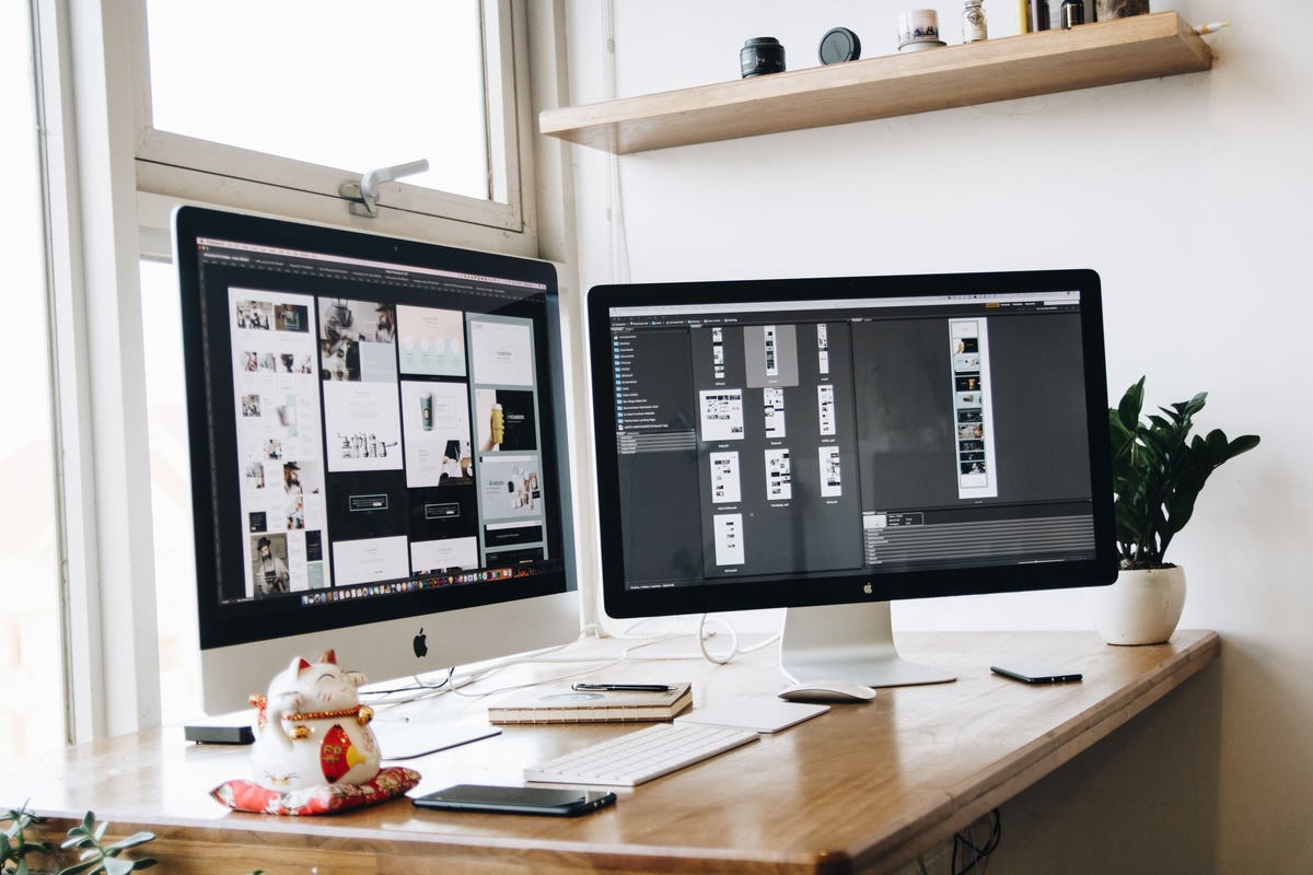 Two Apple iMacs on a light wood desk by a window, with a keyboard, notebook, plant, and lucky cat figurine.